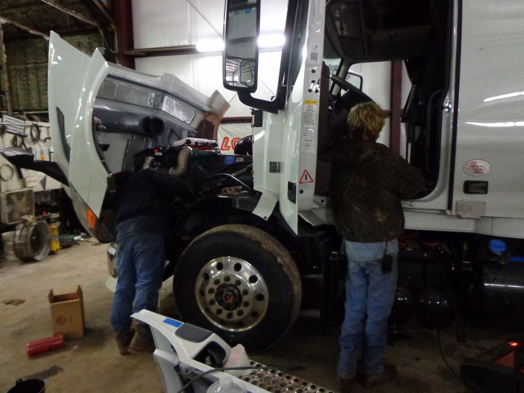 Two mechanics working on a white semi-truck in a garage, hood open and front wheel exposed.