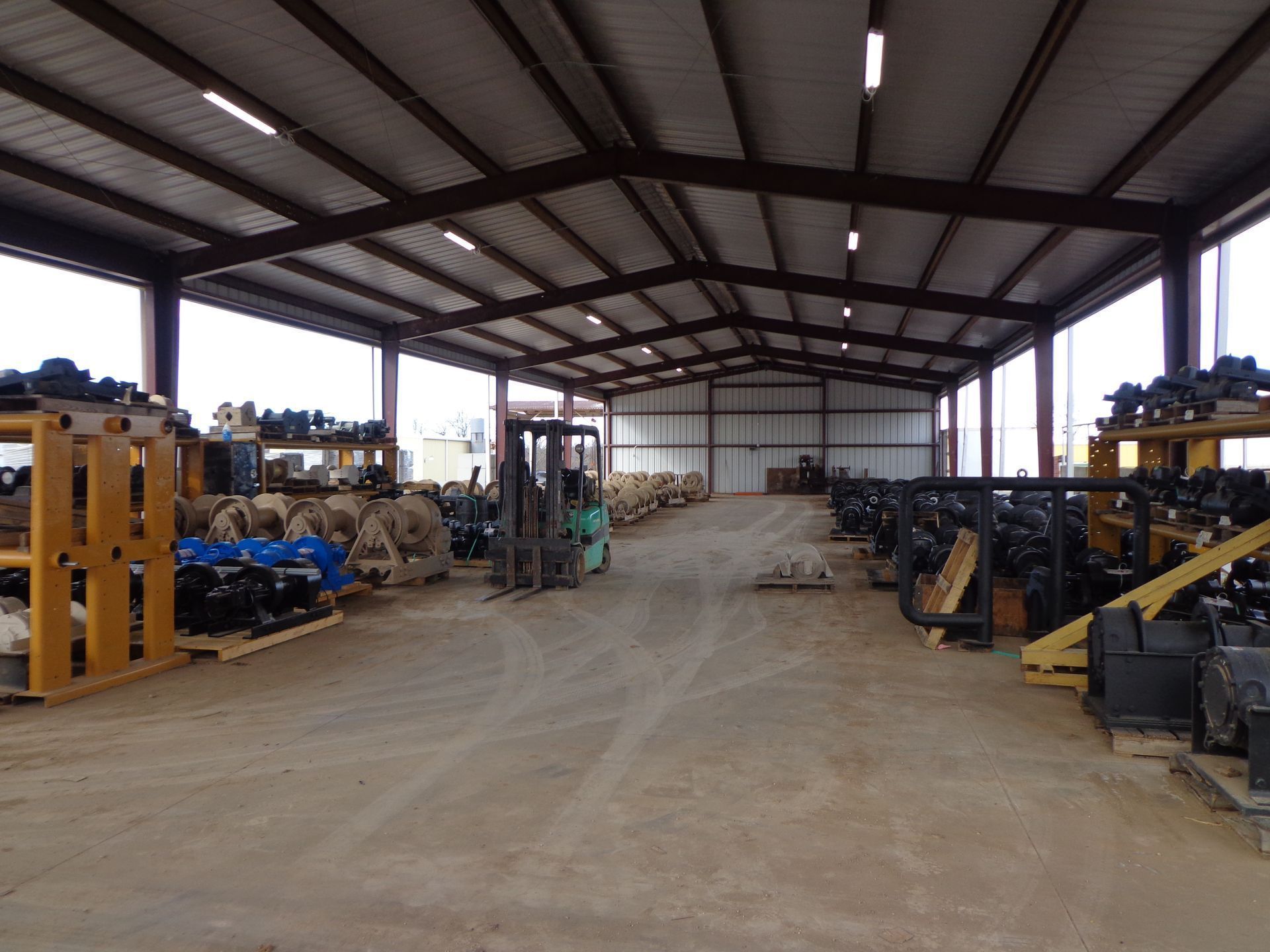 A large warehouse filled with industrial equipment, featuring metal shelving, pallets, and a forklift in the center.