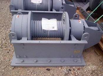 A grey industrial winch drum assembly mounted on a sturdy metal frame, positioned on a gravel surface.