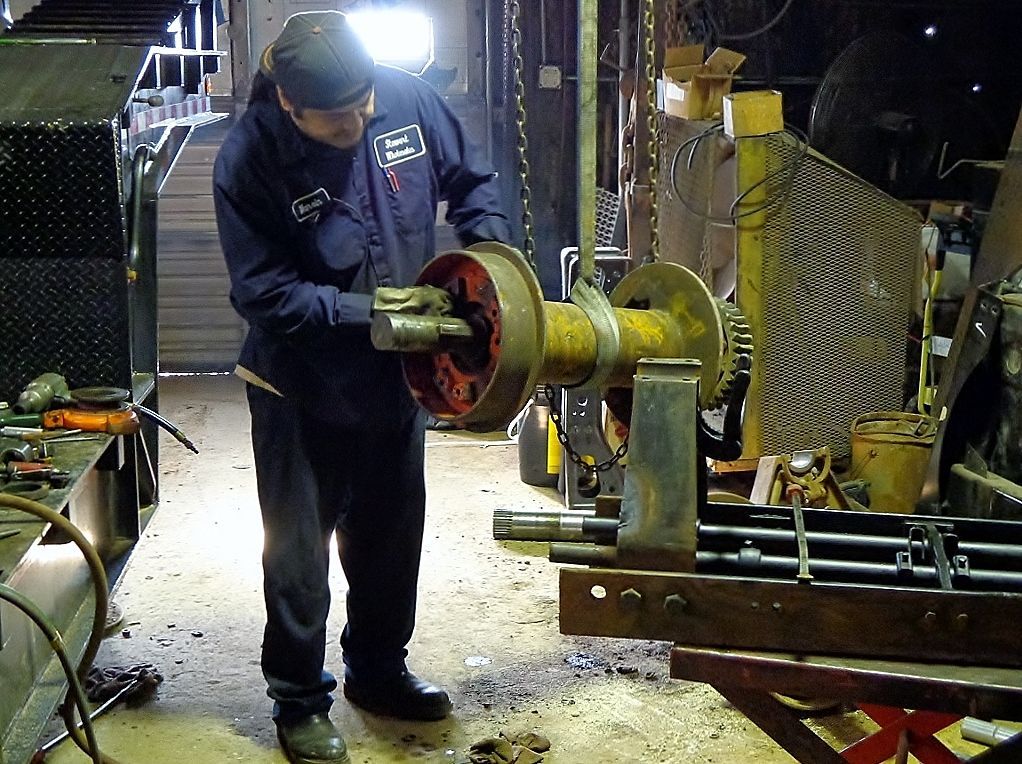Worker handling a large metal spool in an industrial workshop, surrounded by tools and machinery.