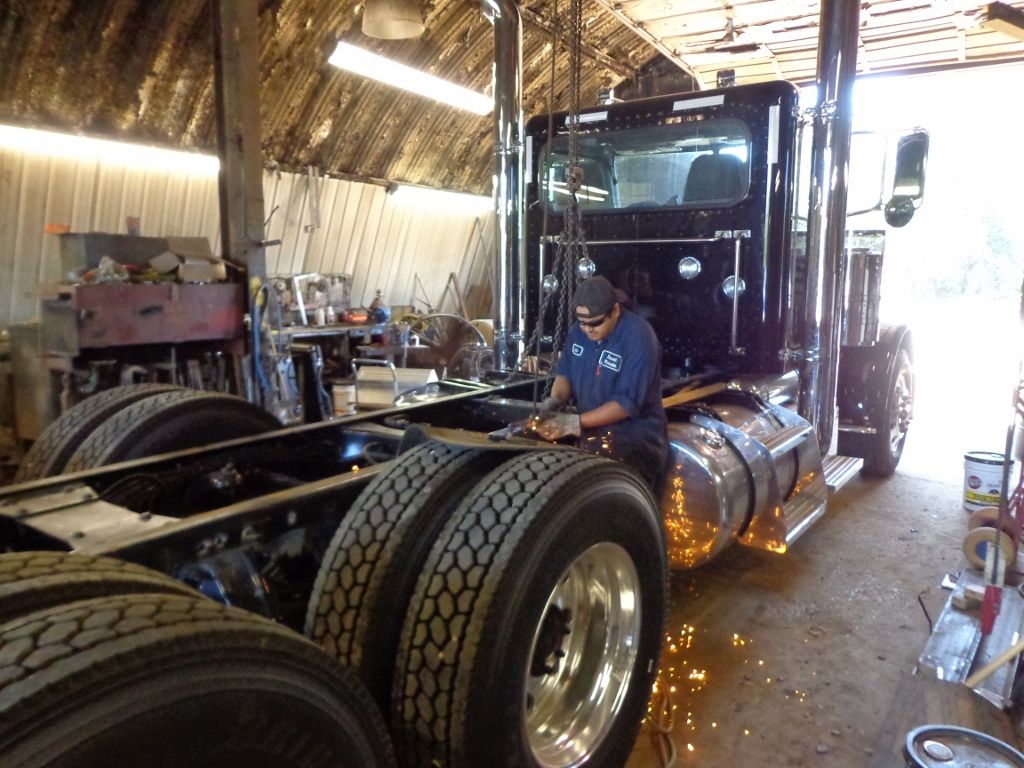 Mechanic welding on a black semi-truck inside a workshop, with sparks flying around the chassis.