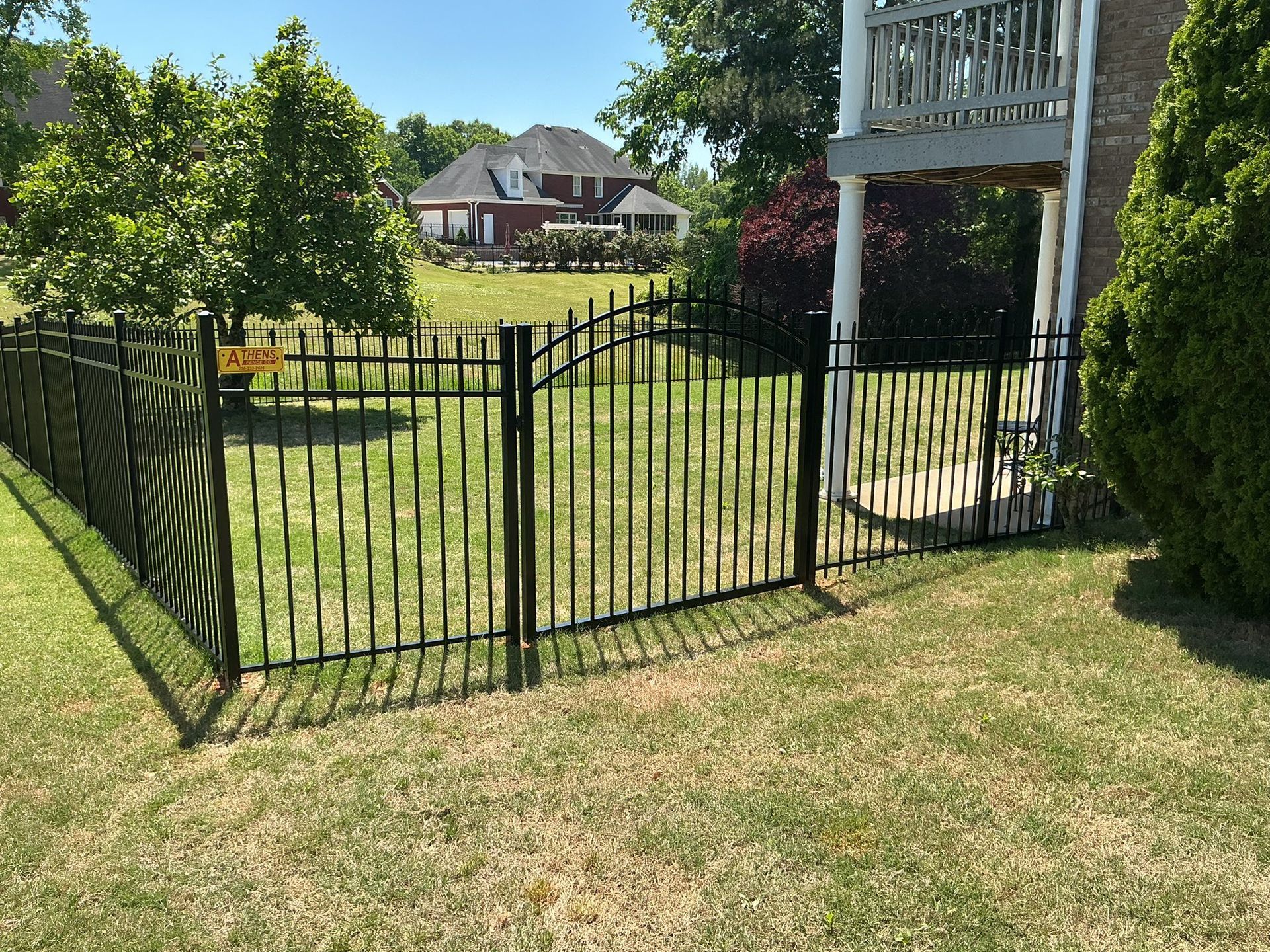 Black metal fence around a grassy yard beside a house and wooden deck, with trees and neighboring homes in back.