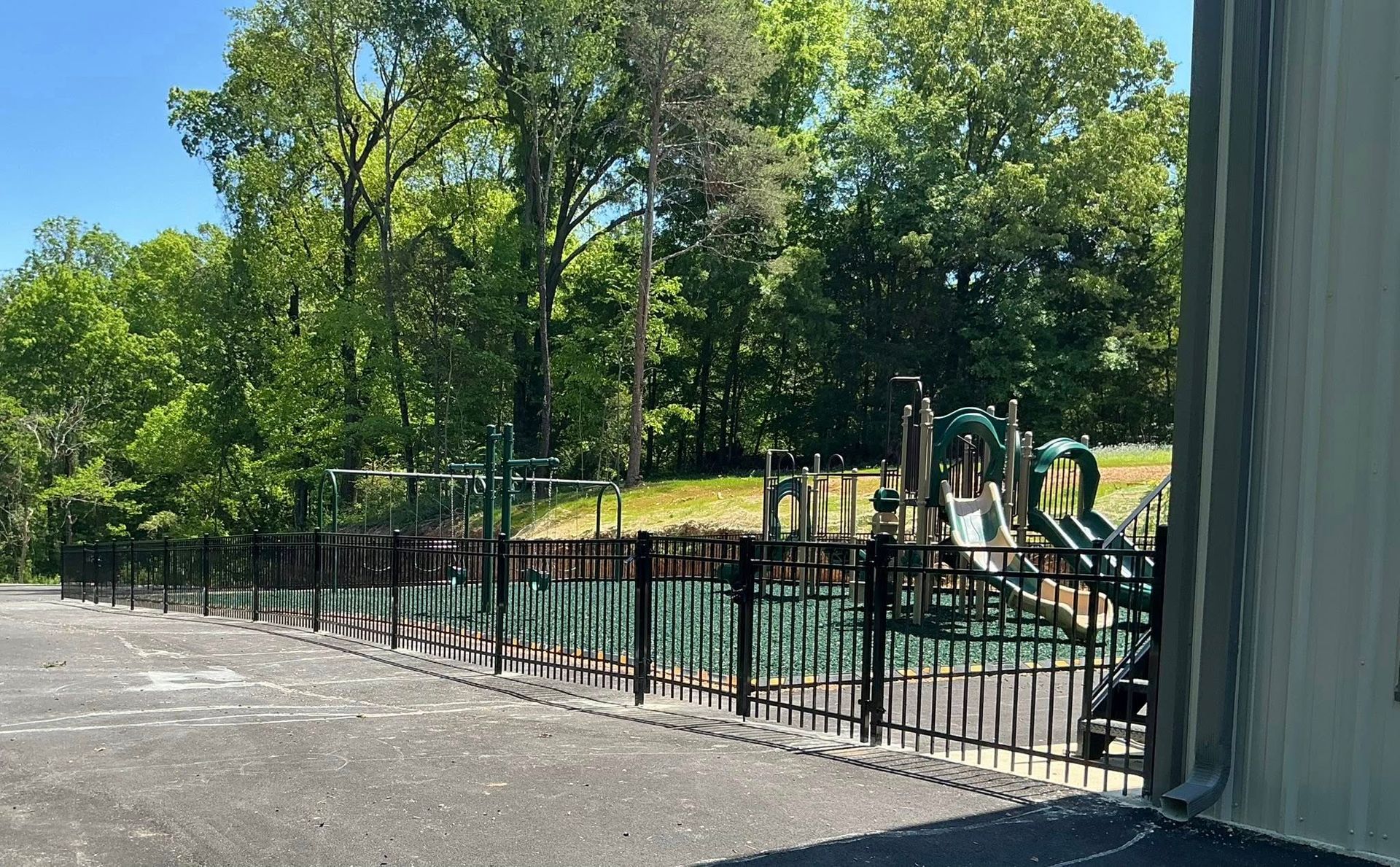 Playground with swing set and fenced area beside trees and a paved path on a sunny day