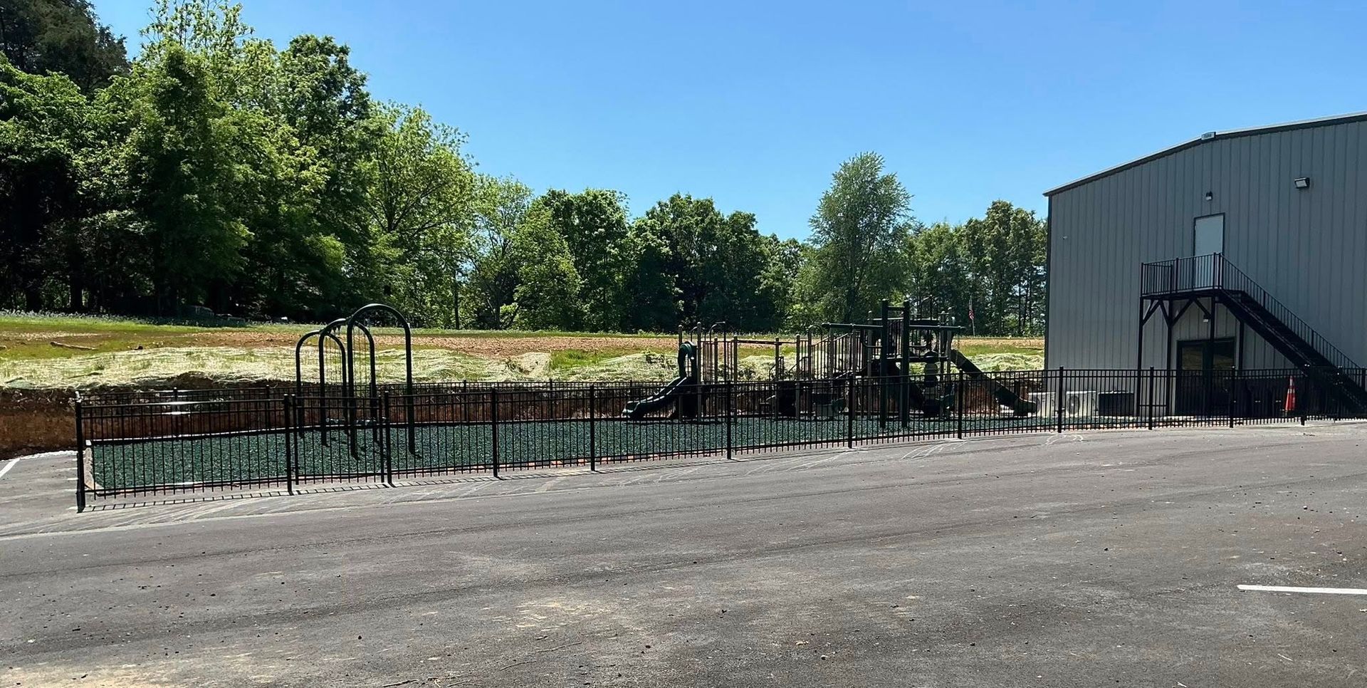 Gravel lot beside a gray building and fenced dirt area, with trees and grass in the background