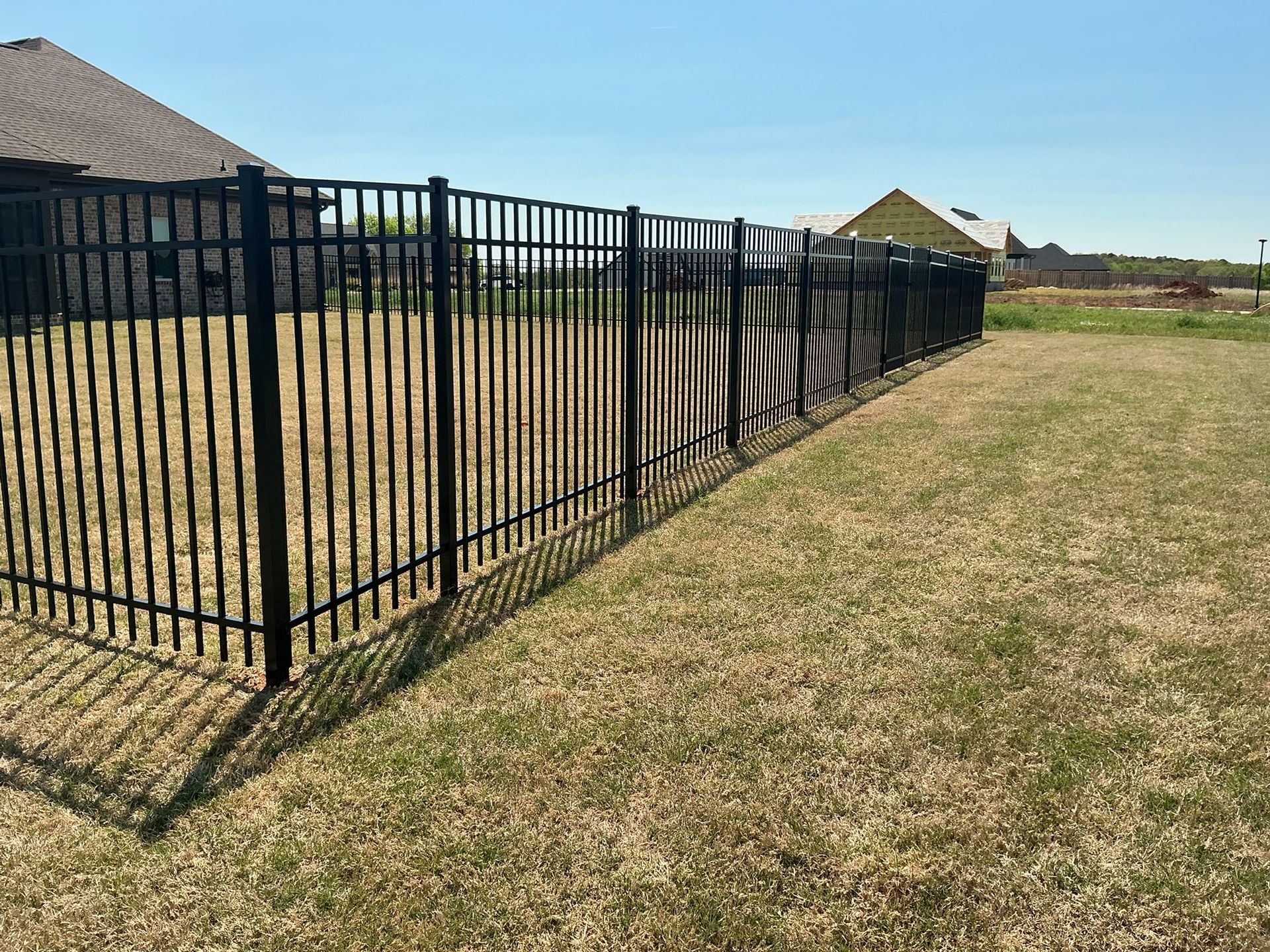 Black metal fence runs along a dry grassy yard beside suburban houses under a clear sky.