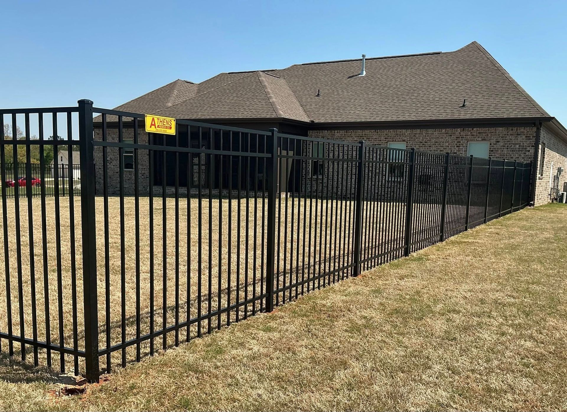Black metal fence around a beige house with a wavy brown roof and dry grass yard under a blue sky