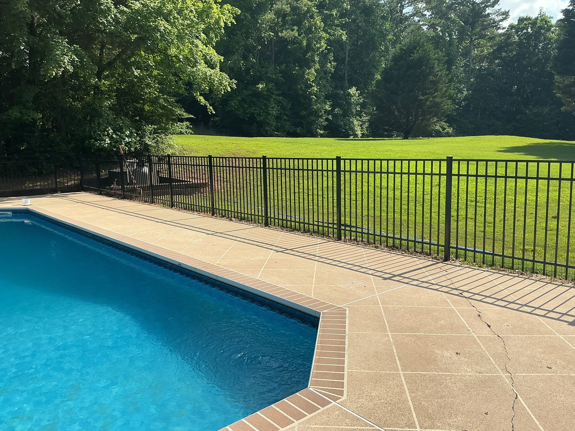 Blue swimming pool beside tan deck and black fence, overlooking a green lawn and trees
