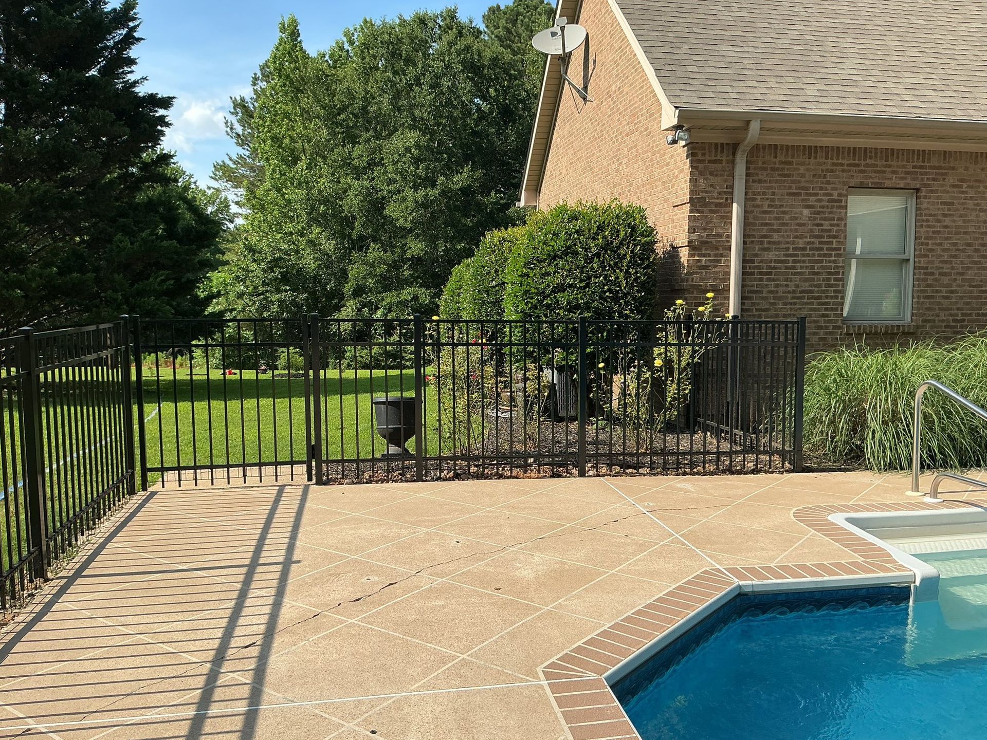 Backyard patio beside a swimming pool, black fence, brick house, and green lawn beyond.