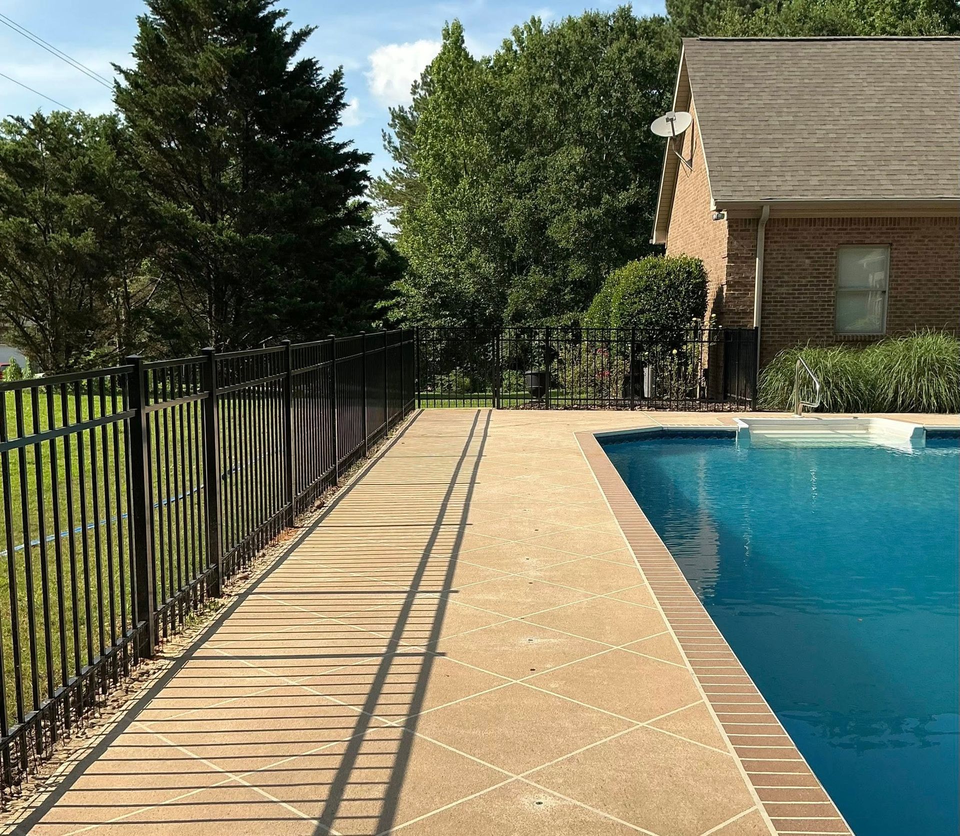 Backyard pool deck with tan tile, black fence, and brick house beside blue water
