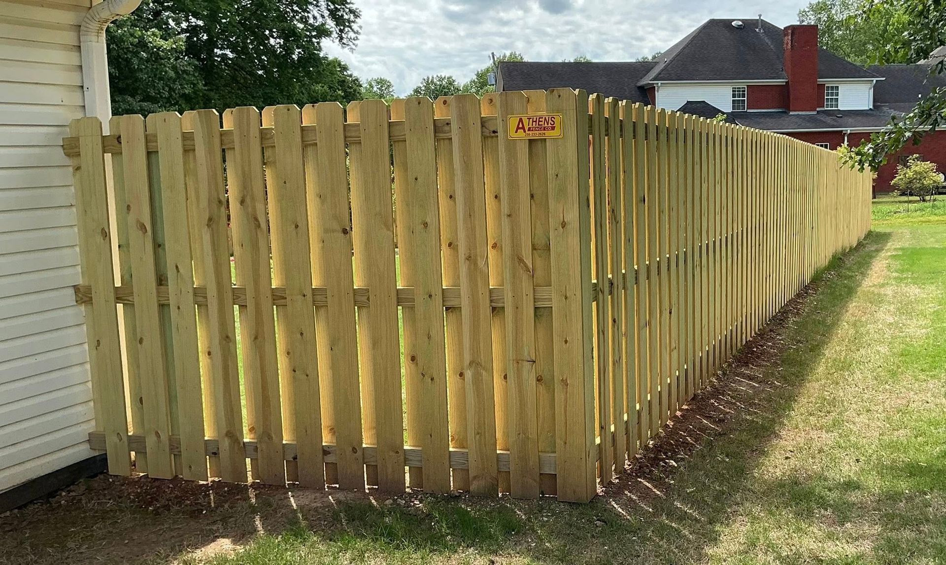 Wooden privacy fence with green grass in front and trees behind under a cloudy sky