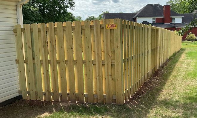 Wooden privacy fence with green grass in front and trees behind under a cloudy sky