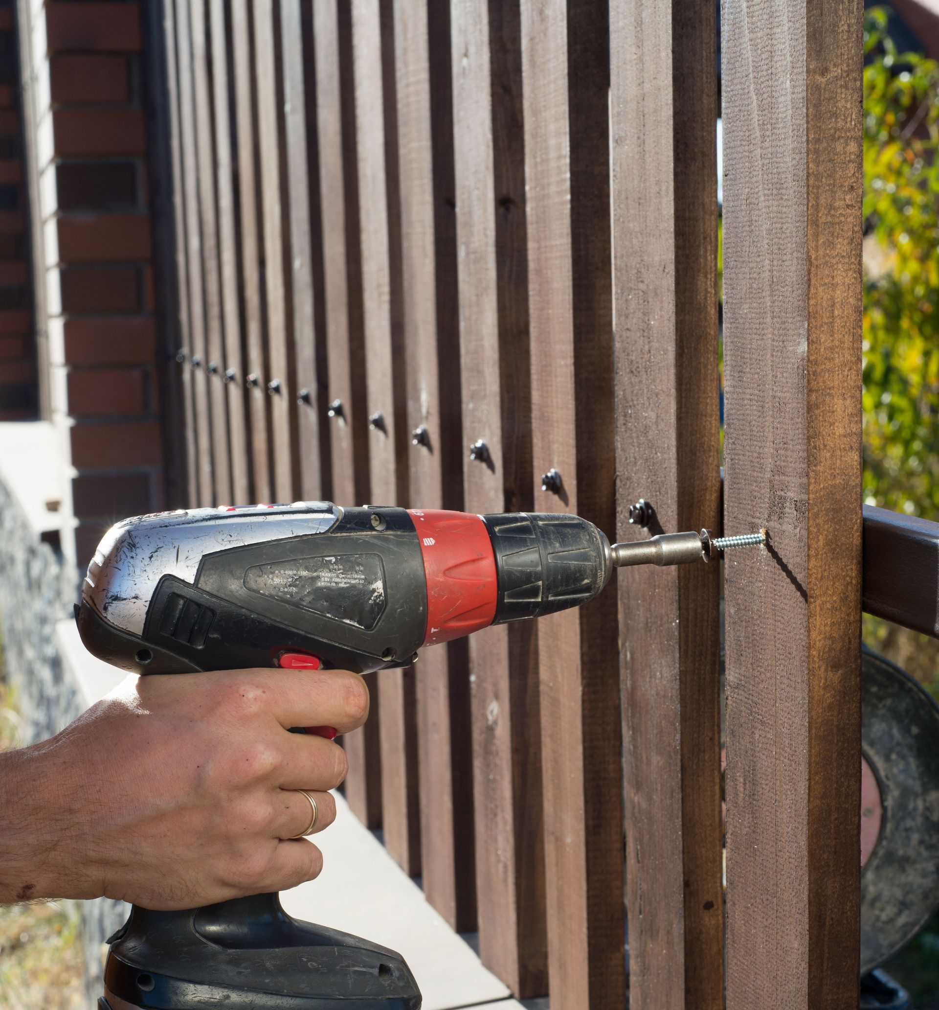 Hand holding a cordless drill driving a screw into a wooden fence rail.