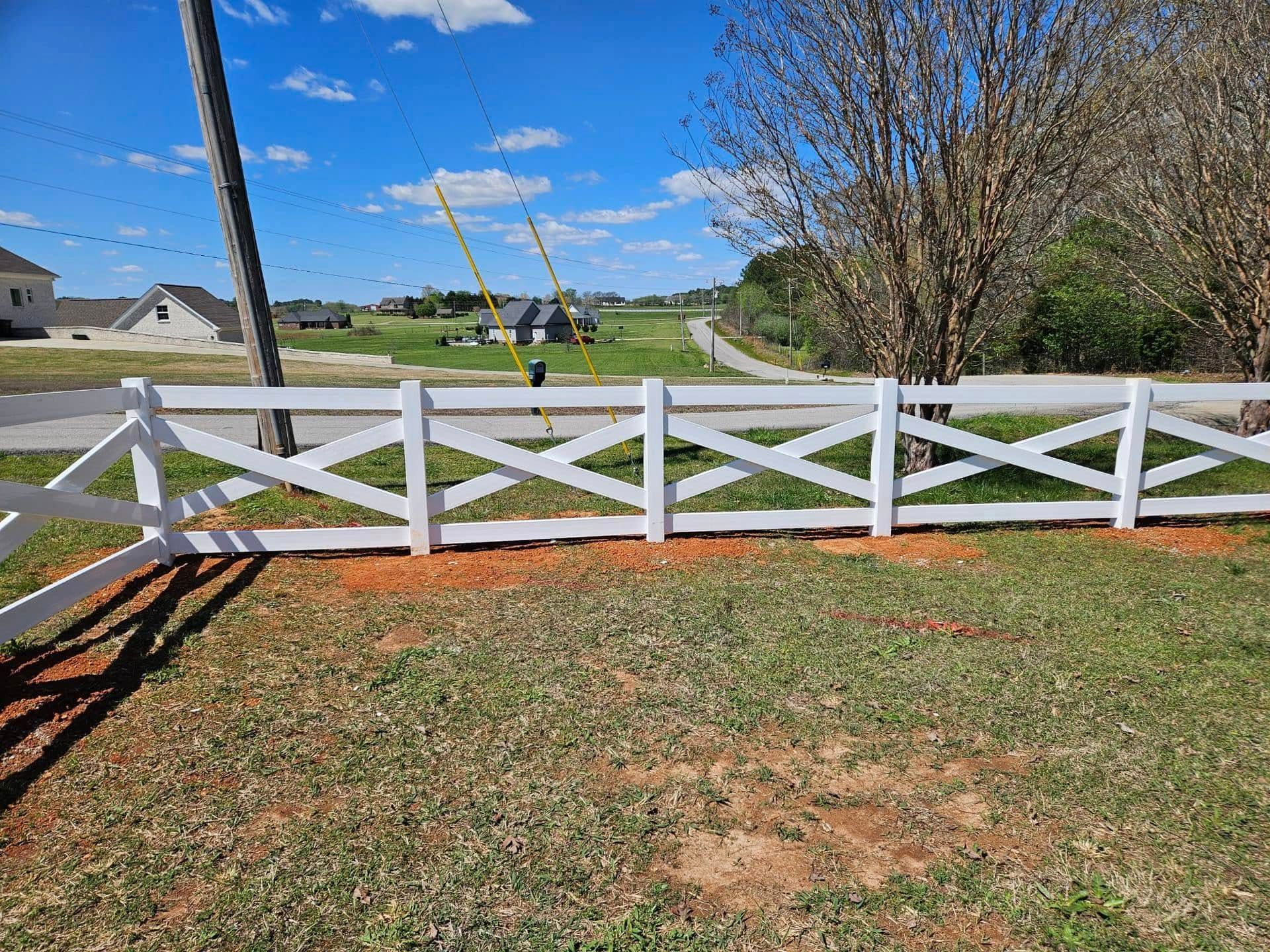 White fence in a grassy yard with a blue sky, tree, and construction crane in the distance.