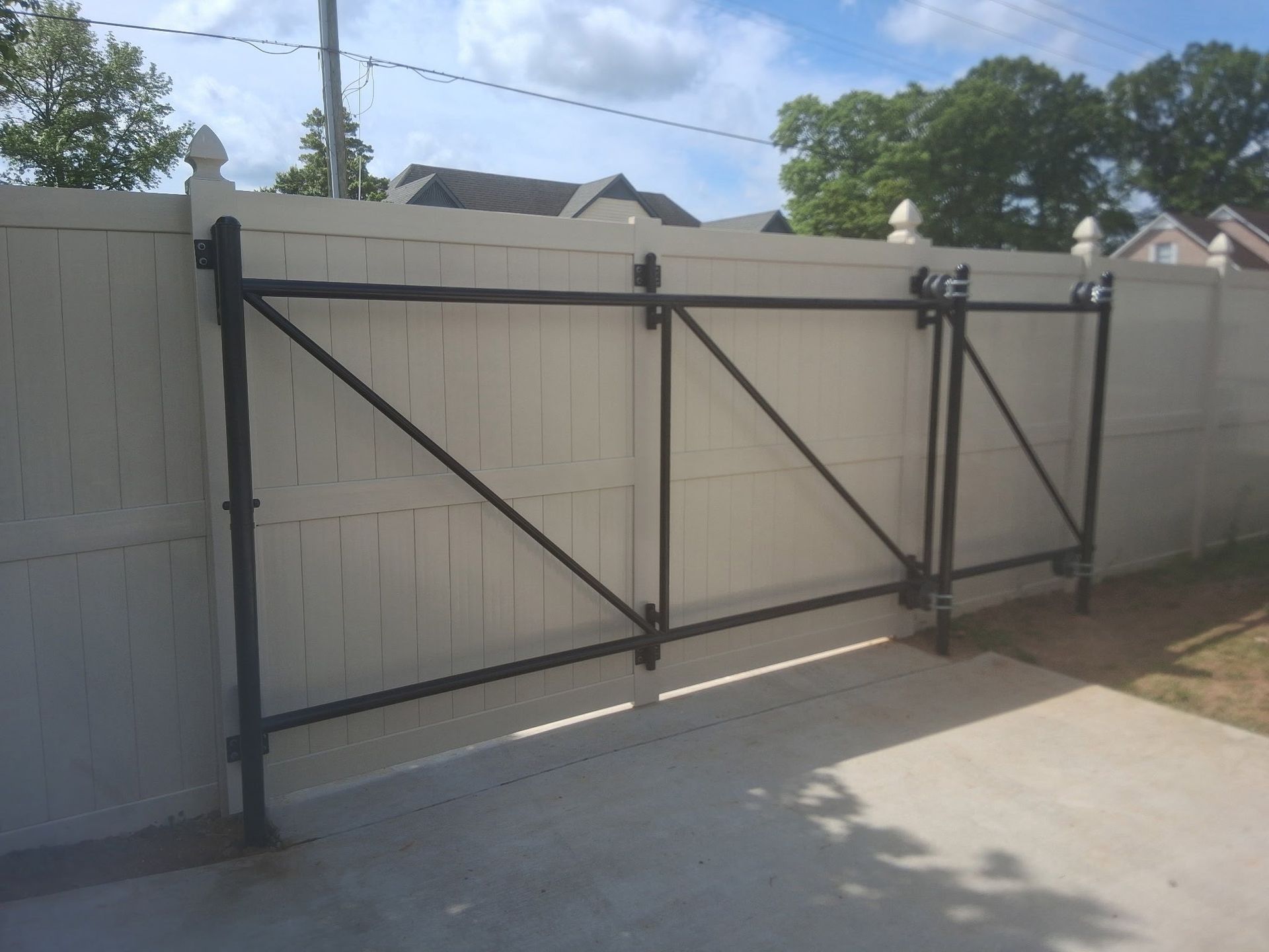Black metal driveway gate with diagonal braces attached to a white fence beside a concrete driveway.