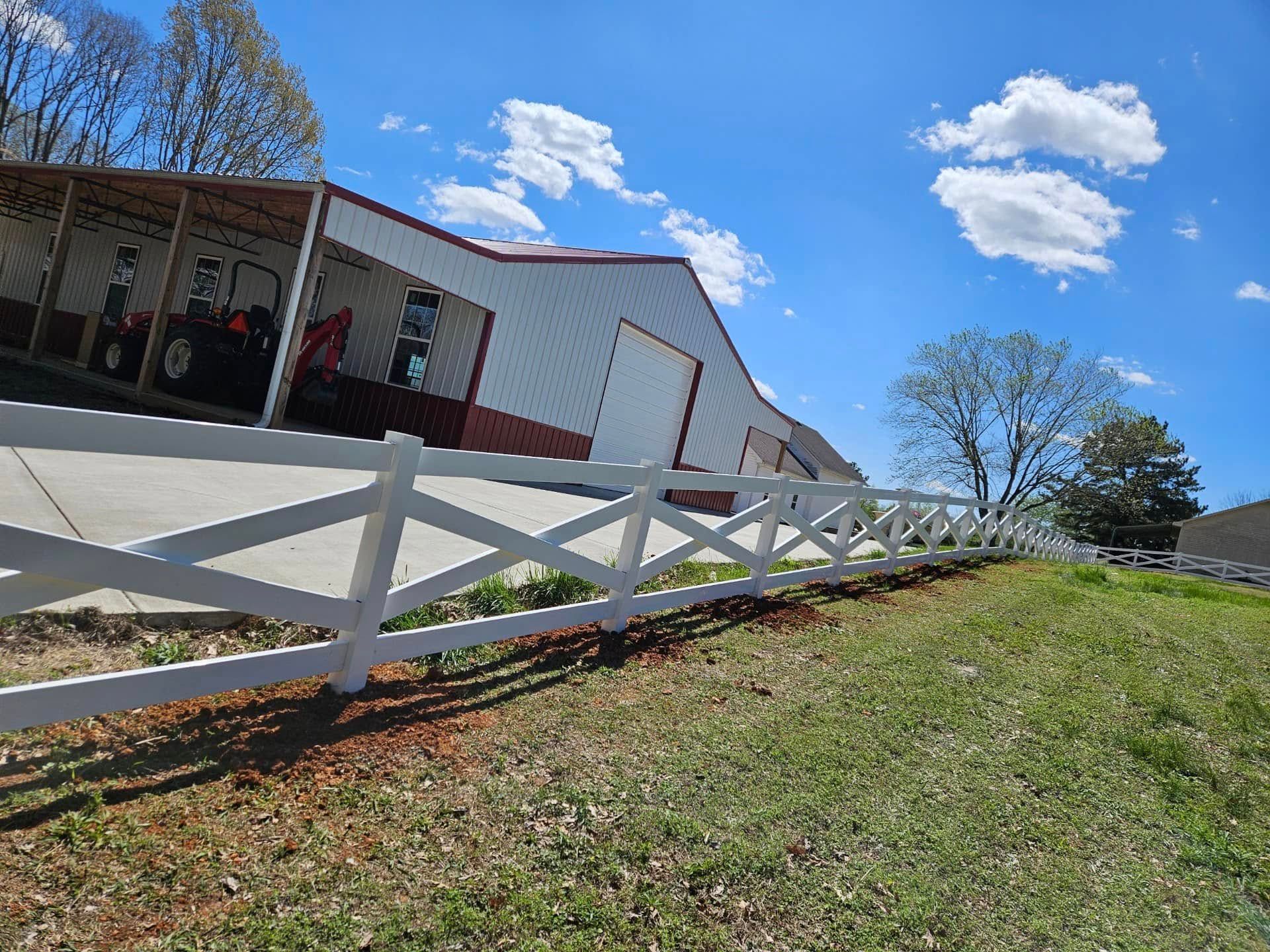 White fence along a grassy slope beside a barn under a bright blue sky with clouds