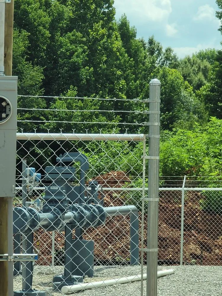 Chain-link fence with industrial piping and valves beside a wooded area on a sunny day