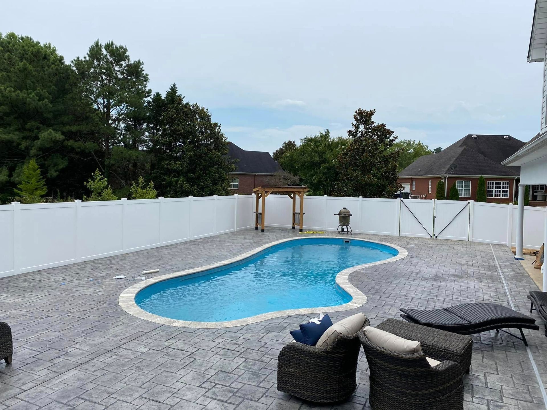 Backyard pool with white fence, gray patio, and lounge chairs under a cloudy sky