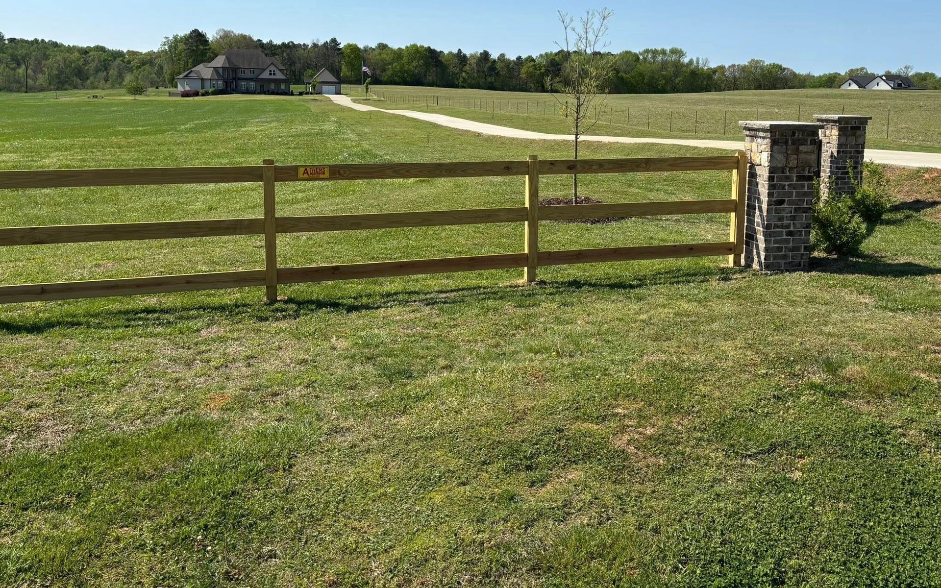 Wooden fence beside a grassy field with a winding path and distant house under a clear sky