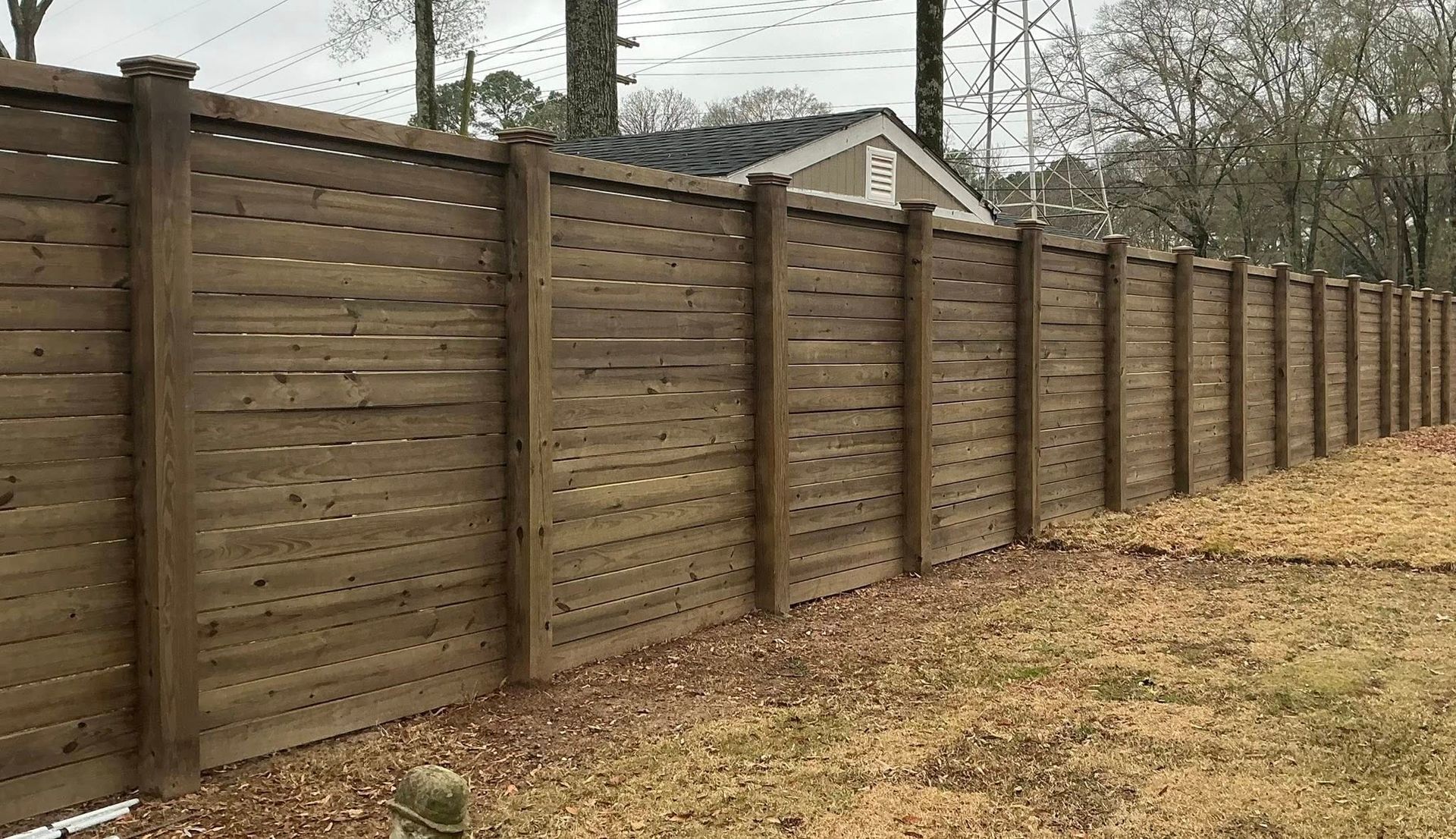 Long wooden privacy fence along a grassy yard, with trees and a small house in the background