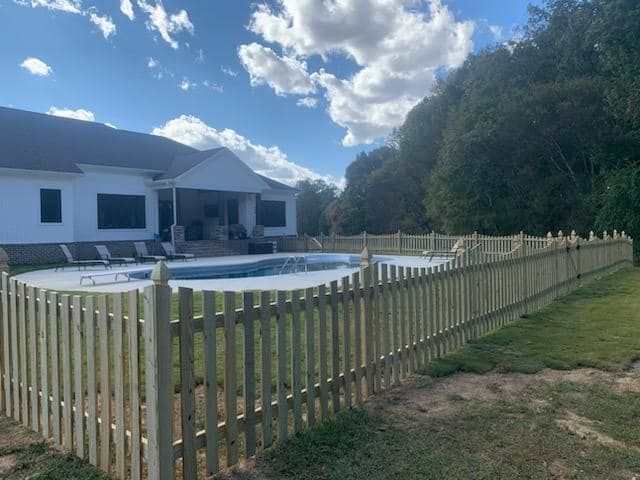 Backyard with above-ground pool, white picket fence, and house under a cloudy blue sky