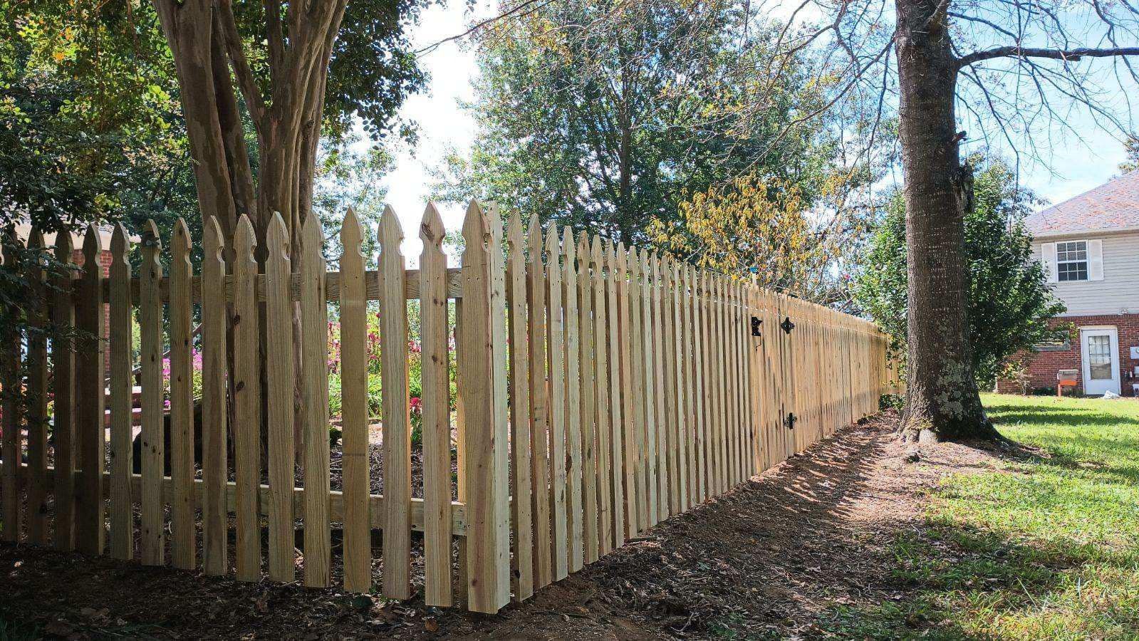 Wooden picket fence beside trees in a leafy yard, with a house in the background.