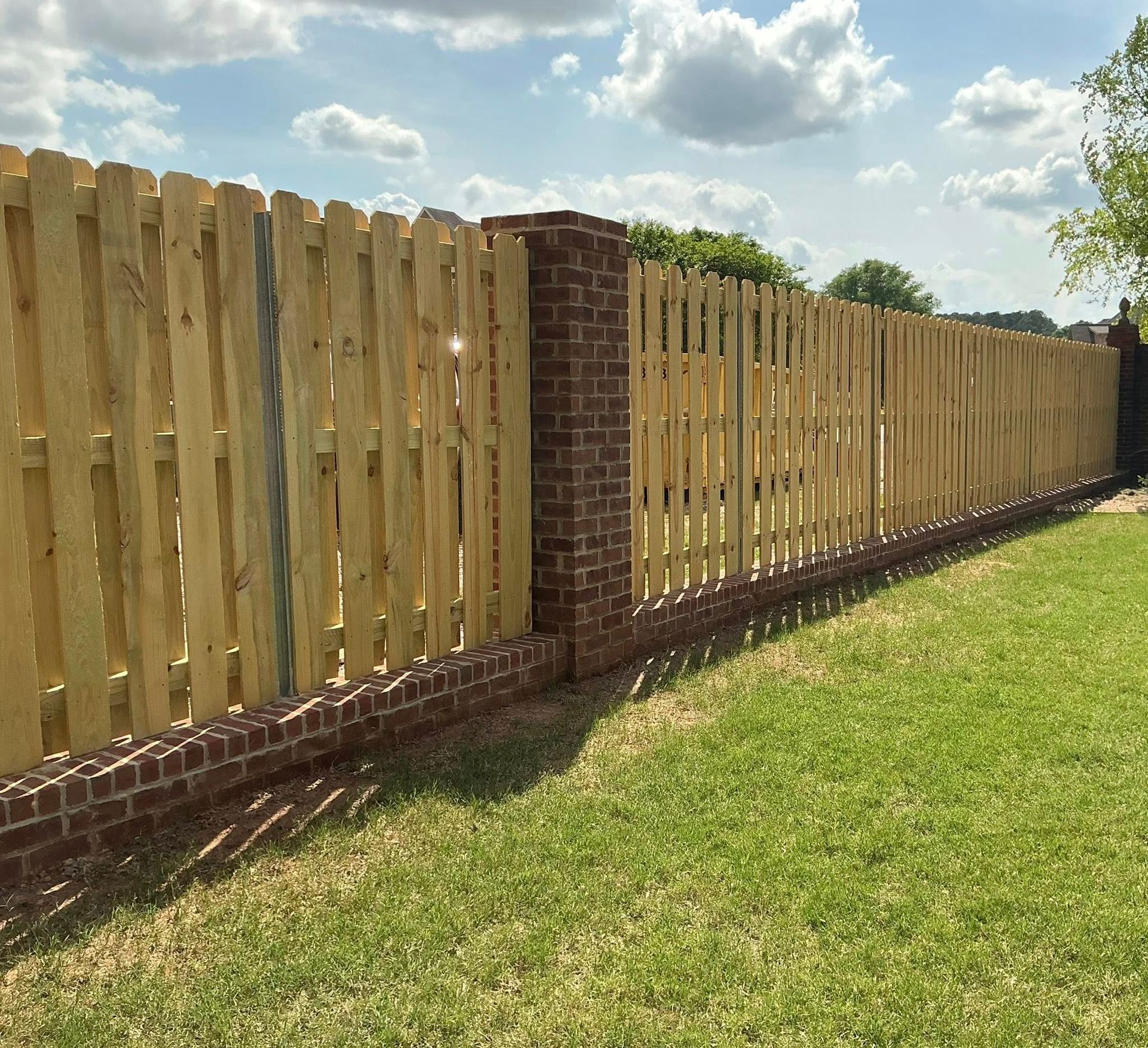 Wooden picket fence with brick columns along a grassy yard under a partly cloudy sky