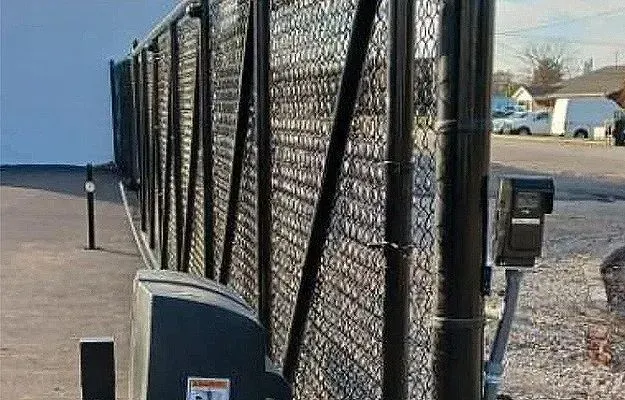 Chain-link fence along a waterfront with utility boxes and a distant shoreline under a cloudy sky