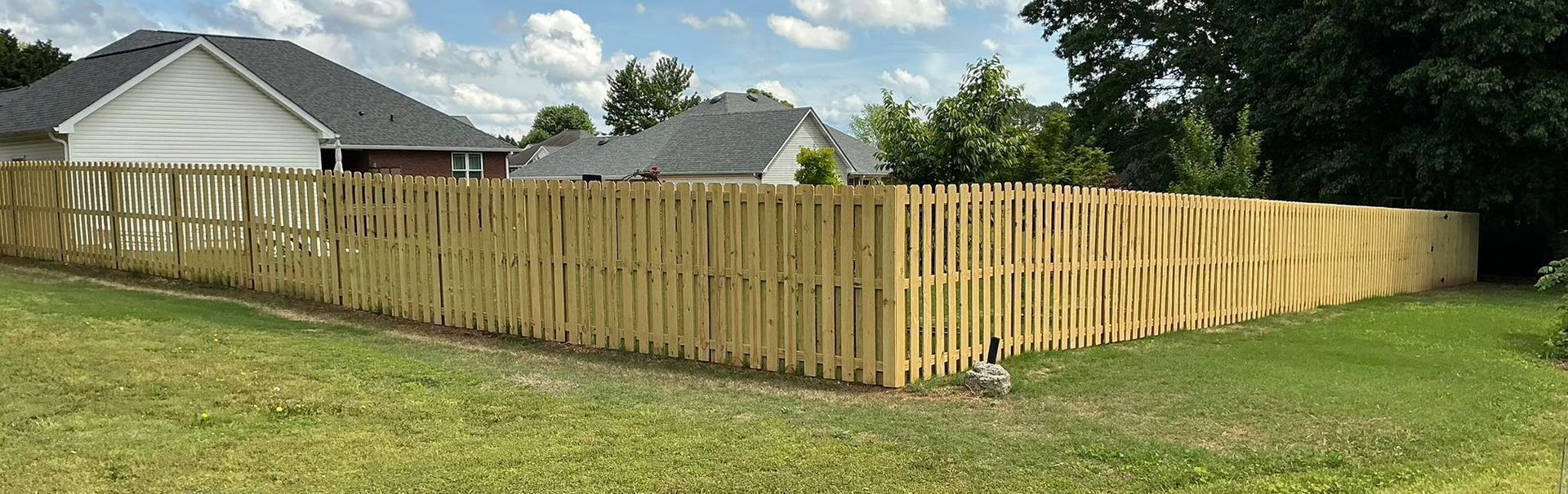 Wooden privacy fence along a grassy yard beside suburban houses under a partly cloudy sky