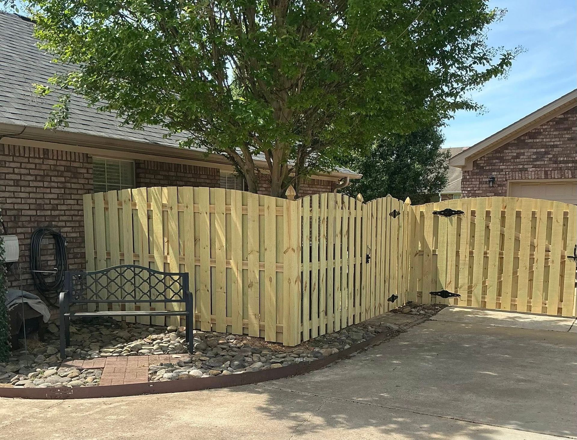 Wooden privacy fence beside a brick house, with a tree and small metal bench in a gravel yard.
