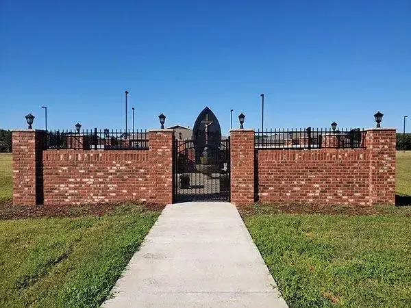 Brick memorial entrance with black iron gate at the end of a concrete path, under a clear blue sky