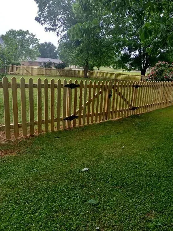 Wooden picket fence with a gate beside a grassy yard and trees on an overcast day