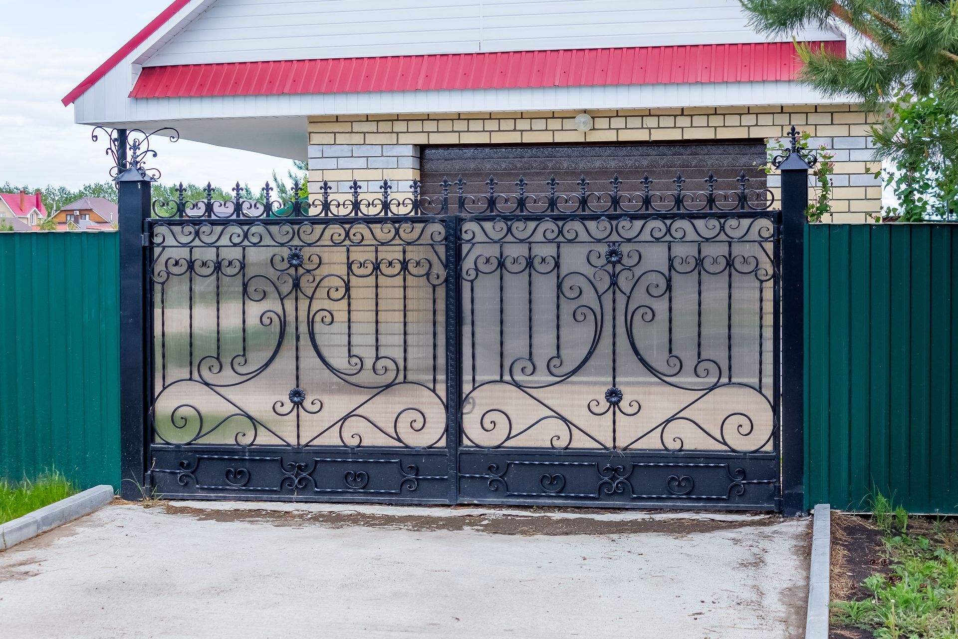 Black wrought-iron gate in front of a house with a red roof and green fence panels.