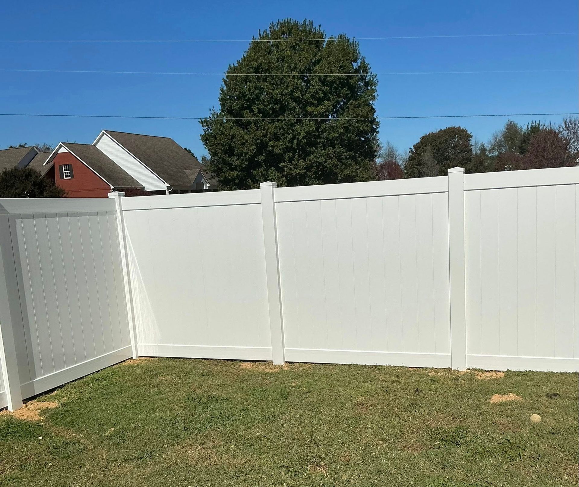 White backyard fence with a gate, lawn, and houses and trees in the background under a blue sky