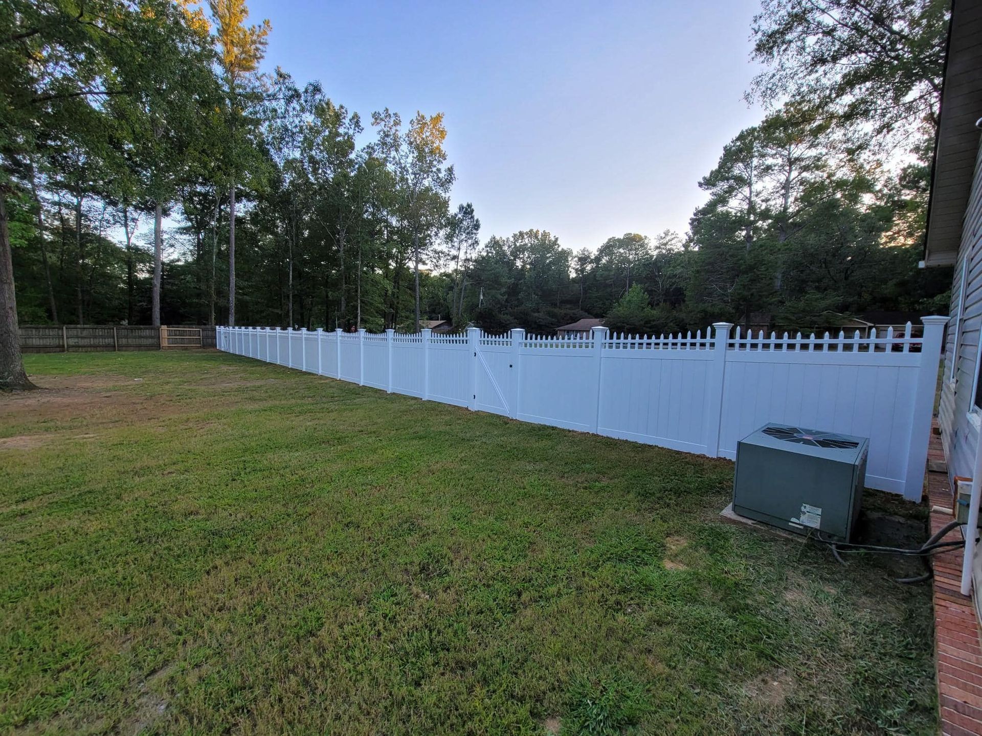White picket fence along a grassy yard at dusk, bordered by trees and a brick corner on the right.