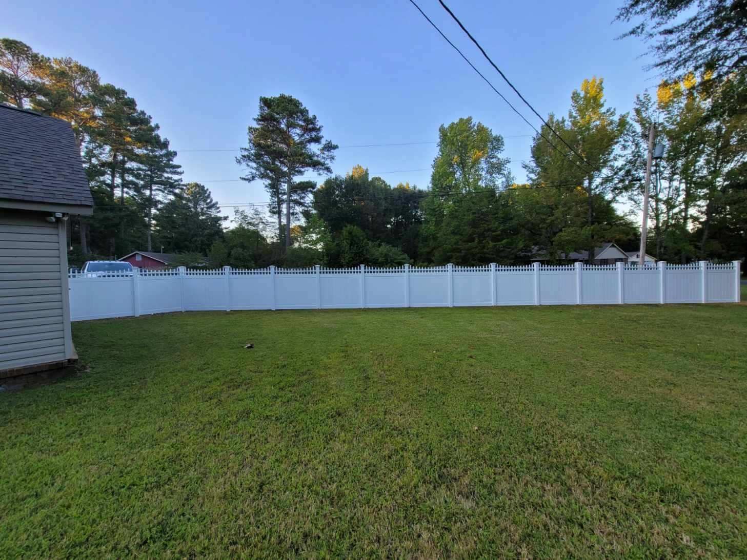 Backyard lawn with white vinyl fence, trees, and a house at the left edge at dusk
