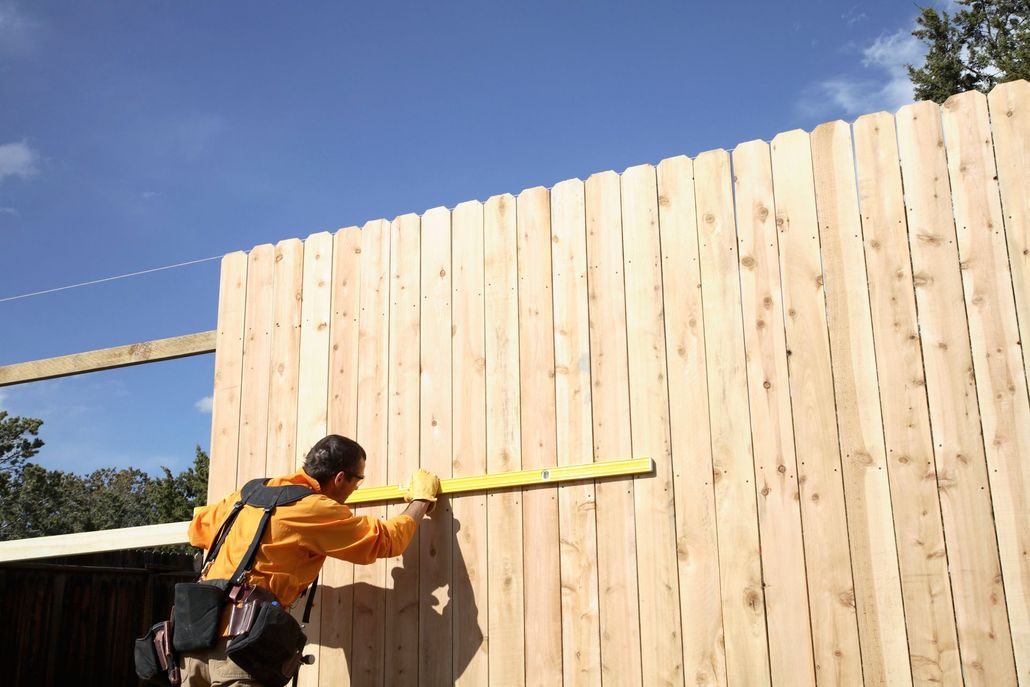 Worker in orange shirt measuring a tall wooden fence with a tape measure under a blue sky