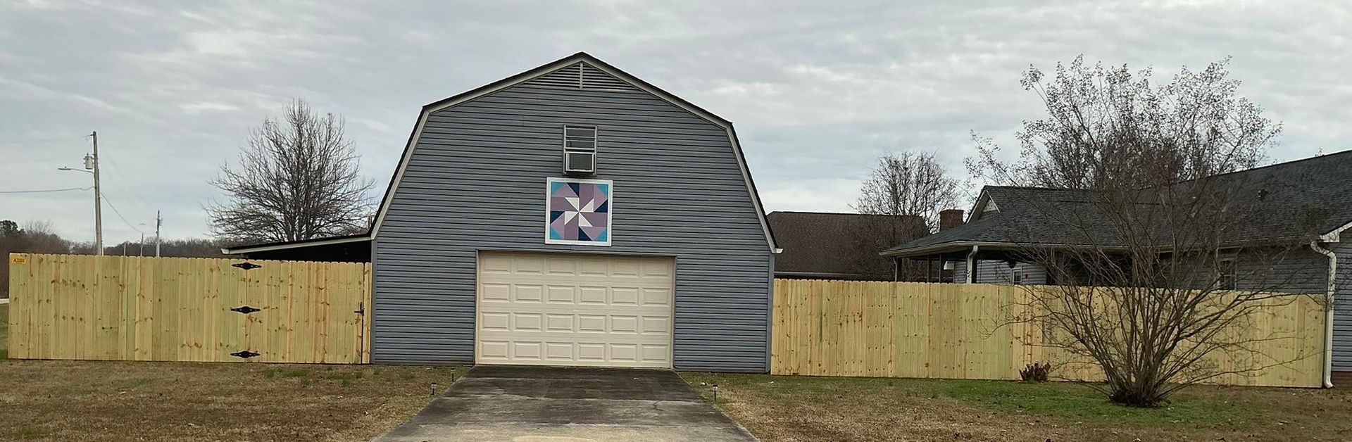 Gray barn-like building with a large garage door, bordered by wooden fences under a cloudy sky