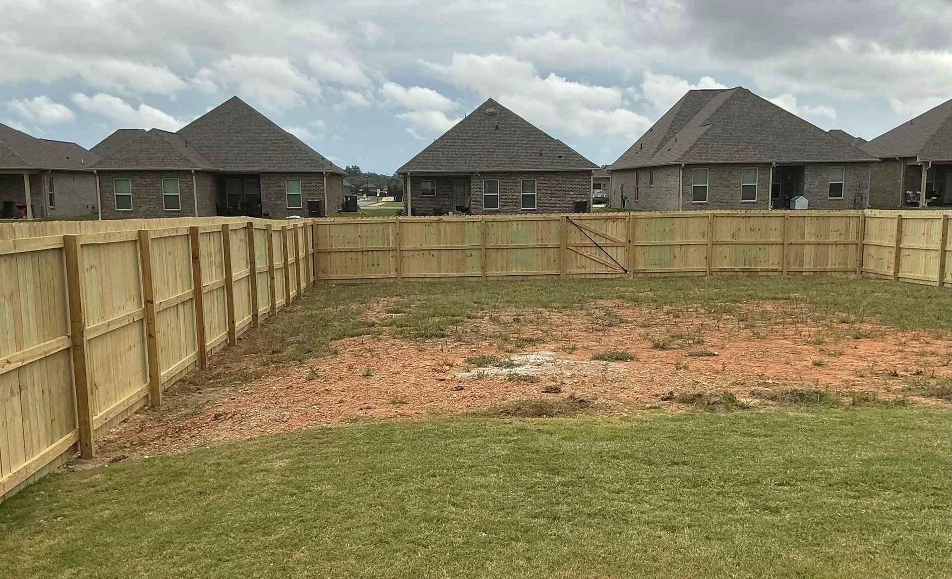 Small fenced backyard with bare dirt patch and neighboring houses under cloudy sky
