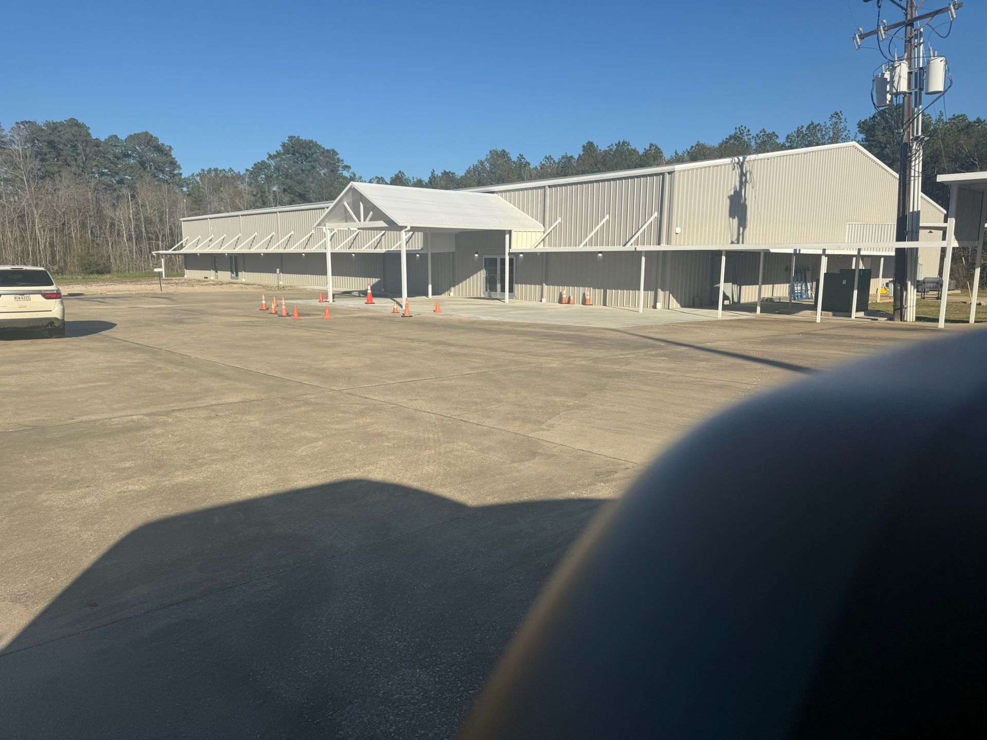 A commercial building under construction on a sunny day. The building is white with a covered entrance, and there's a car and traffic cones in the gravel lot.