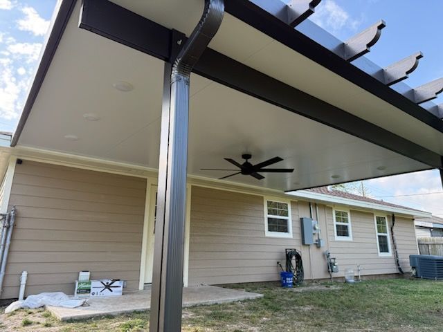 Brown-roofed patio extension attached to a tan house. Includes a ceiling fan, gutter, and part of a pergola. Over a grassy backyard.