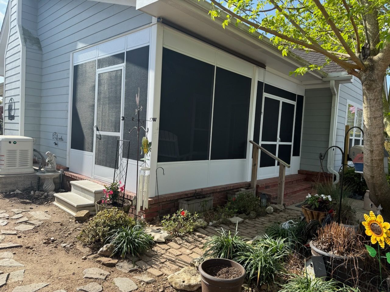 A screened-in porch attached to a light blue house. There are several entrances, stairs, and surrounding flower beds.
