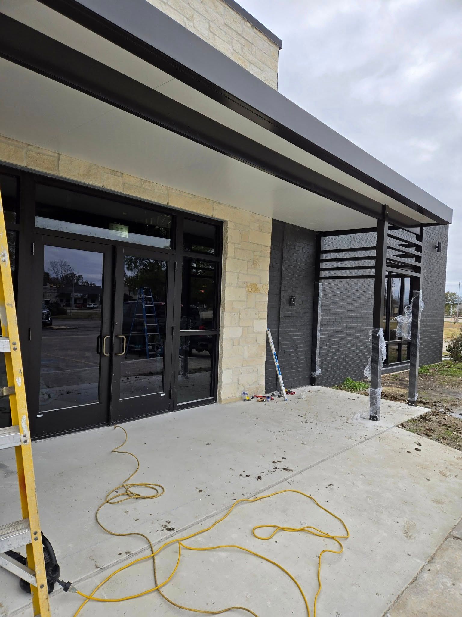 Exterior view of a building entrance with black-framed doors, a concrete porch, and a dark gray awning. A ladder and electrical cord are present.