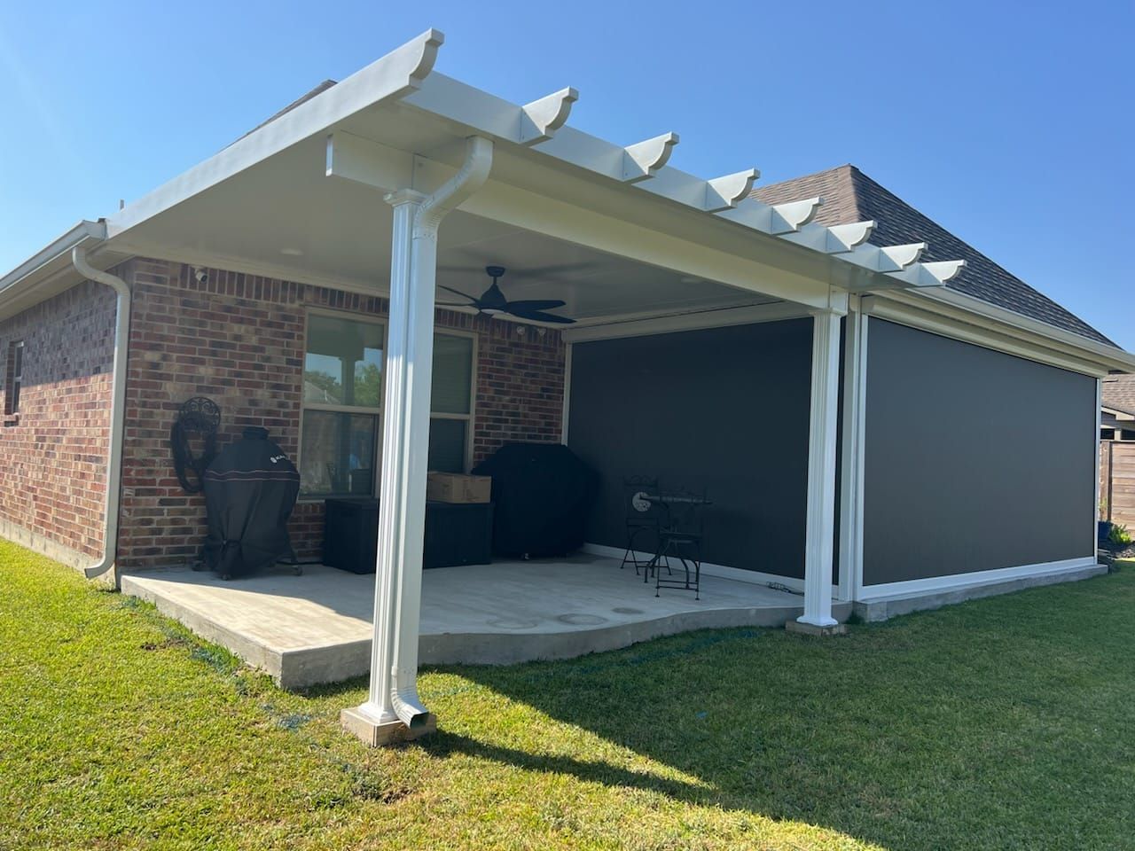 A patio with a white-columned cover and retractable shades, next to a brick house. Green grass surrounds the patio.