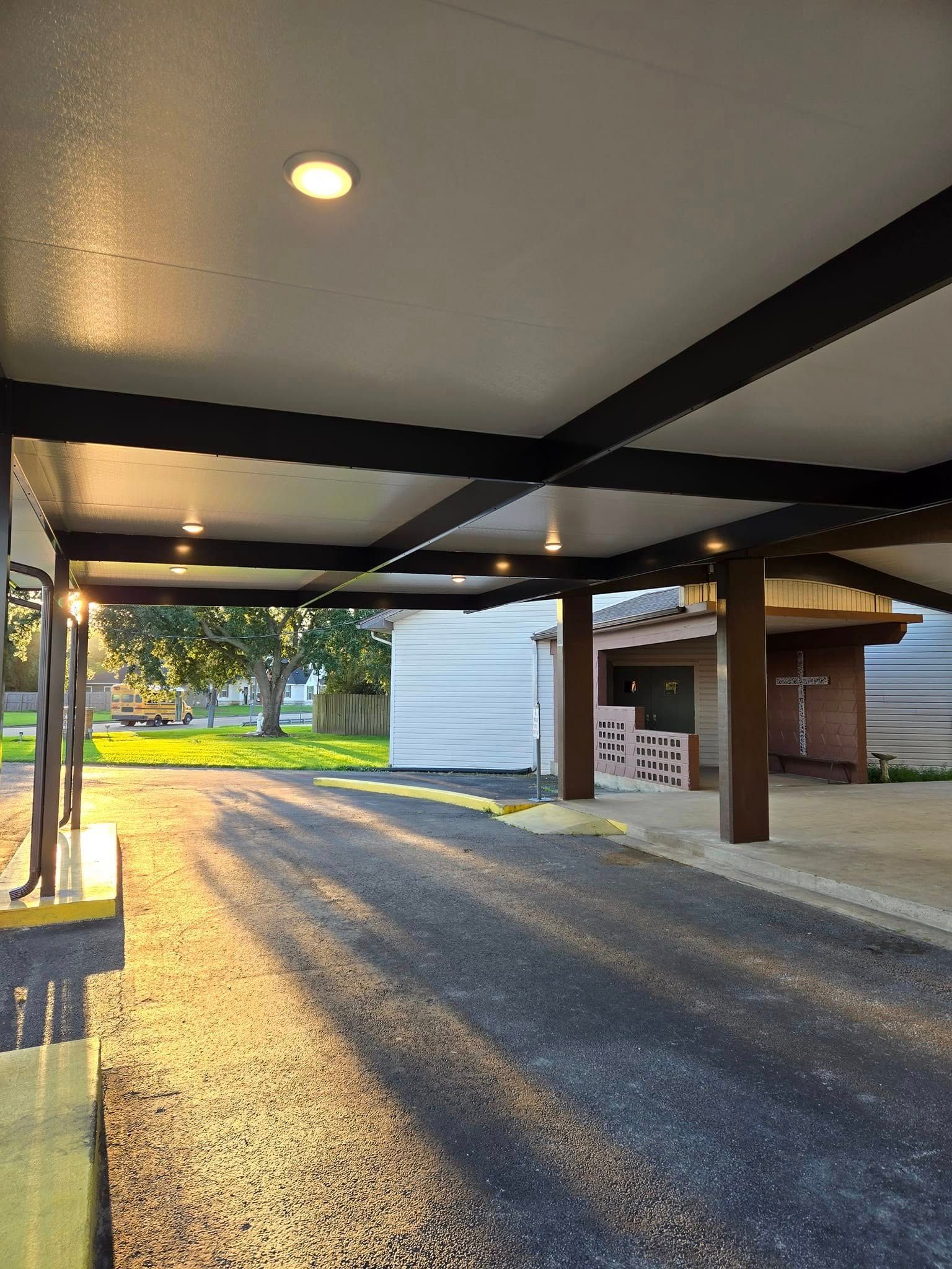 Covered driveway with overhead lights, looking toward a building and a sunny yard. Asphalt, dark beams, and brown support columns are visible.