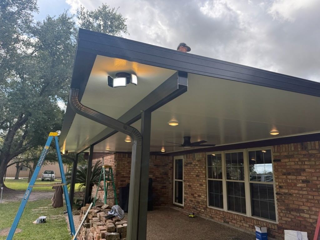 Patio cover attached to a brick house, a person on the roof, gutter and recessed lights are visible.
