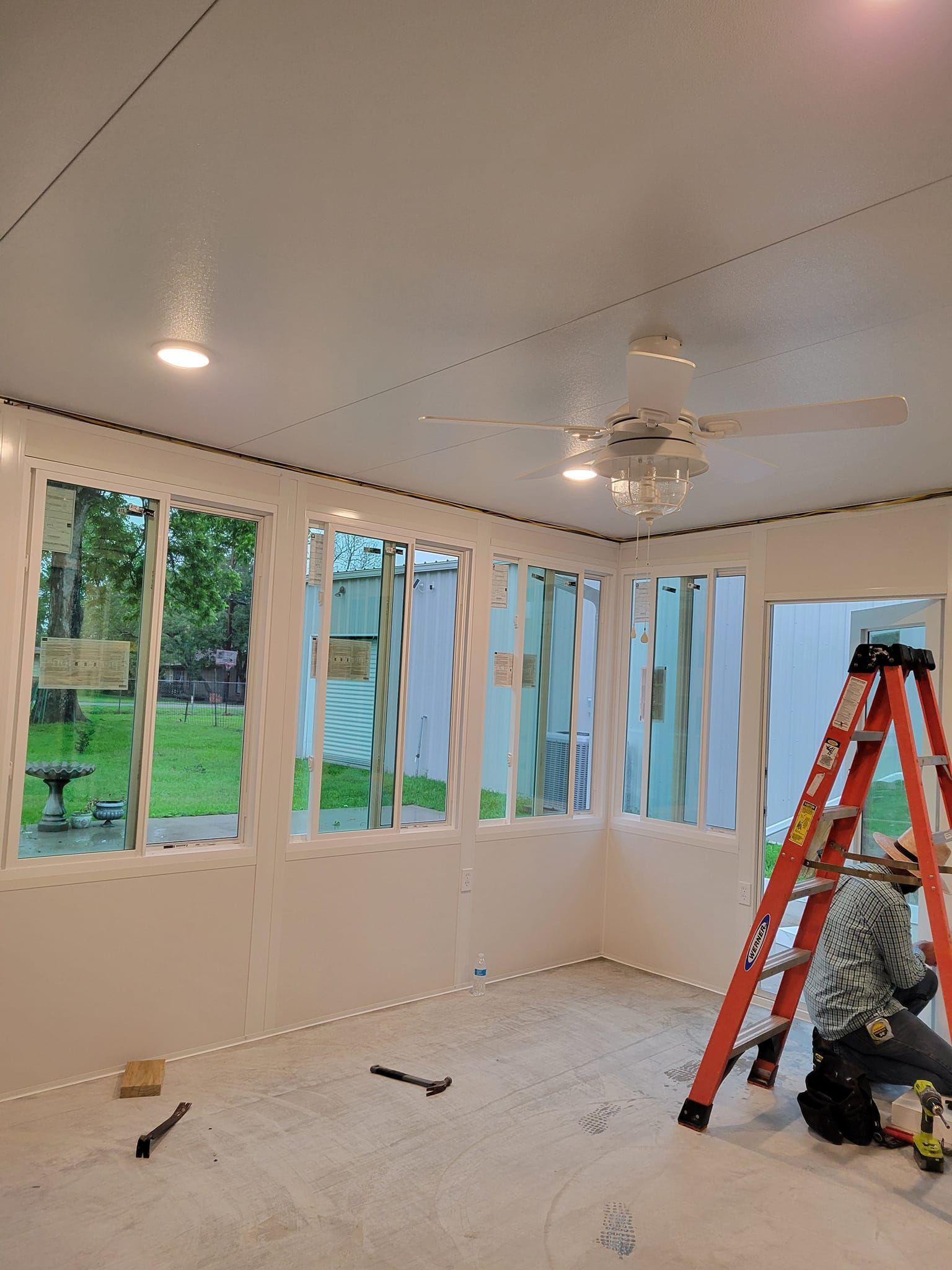 Interior of a room with white walls and windows. A ladder and a person are on the right side. The room is under construction.