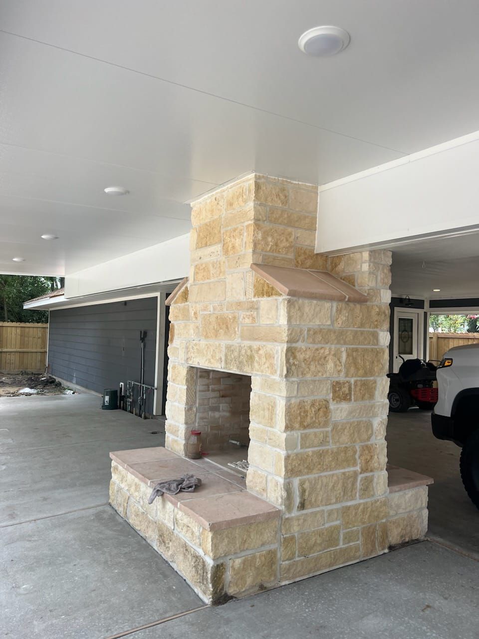 A stone fireplace under a covered outdoor area. The fireplace is tan and brown, with a gray garage door and white ceiling in the background.