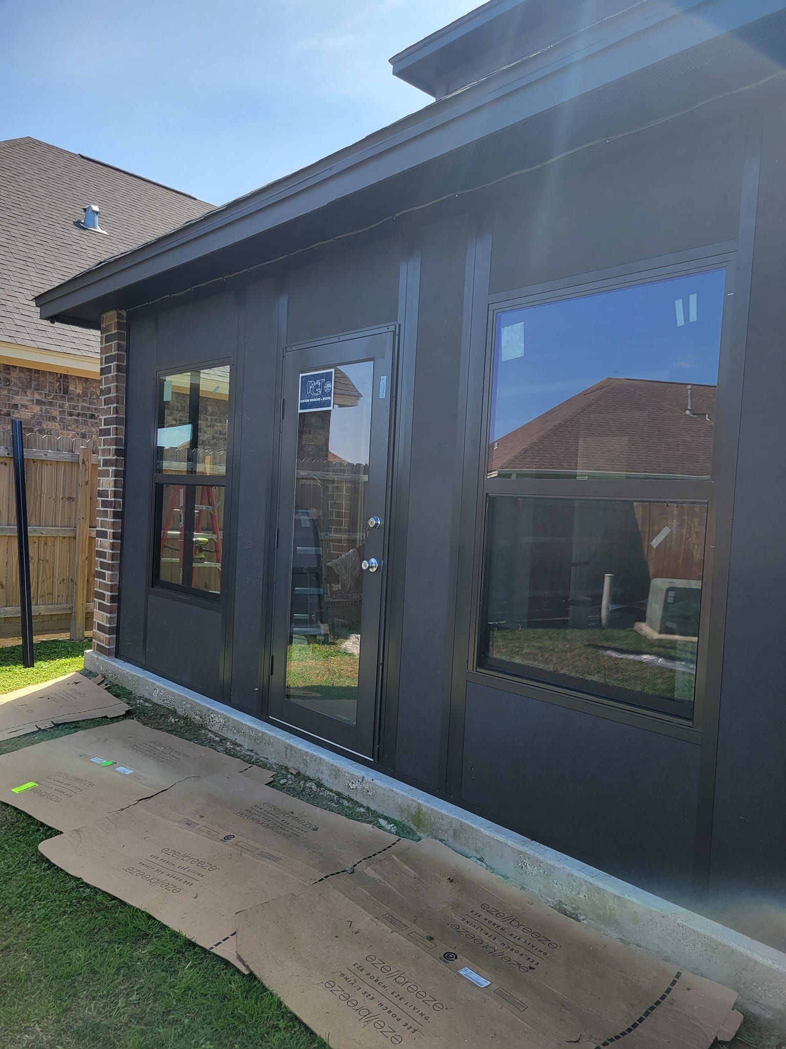 Exterior of a dark-sided building with windows and a door. Cardboard covers the ground in front of the building, and grass surrounds it.