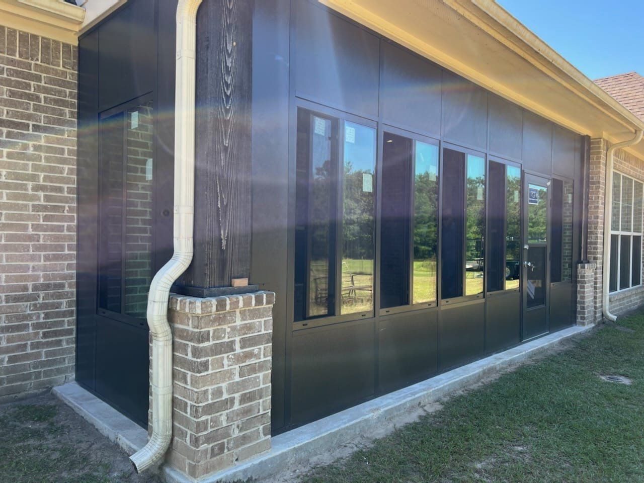 A black-walled sunroom addition with multiple rectangular windows is attached to a brick house. A white rain gutter runs down the side.