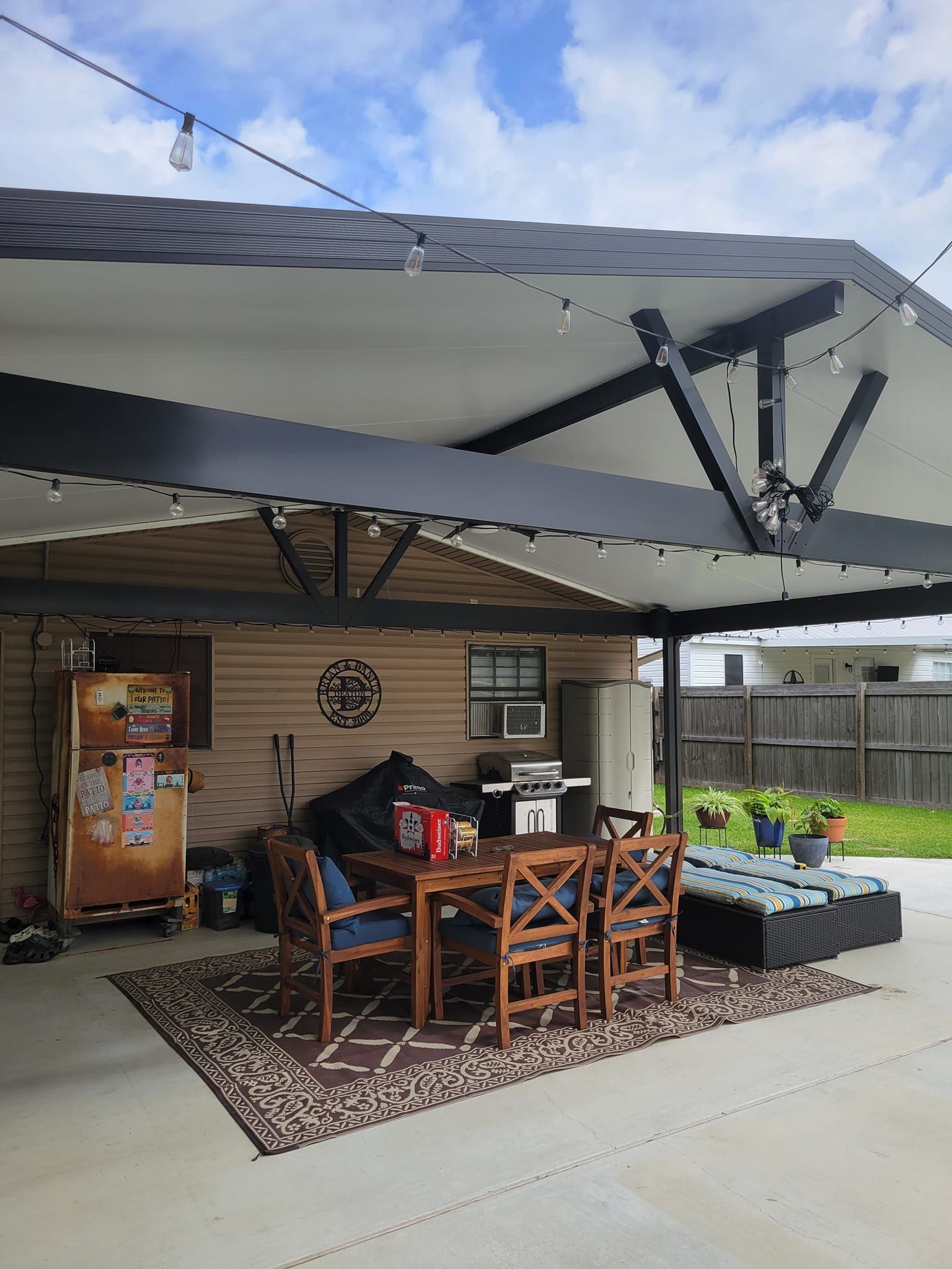 Backyard patio with a dining table and grill under a covered awning. Beige siding, brown rug, and blue chairs are visible.